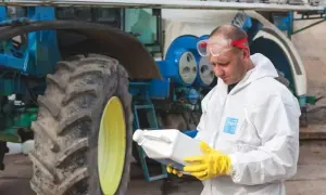 Rolnik w kombinezonie ochronnym i rękawicach trzyma butelkę z preparatem, prawdopodobnie zawierającym difenokonazol, obok traktora.