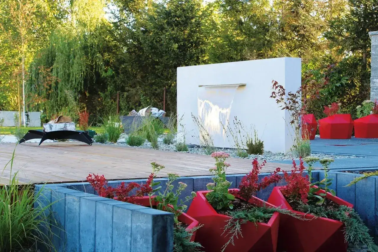 Nowoczesny ogród z małą kaskadą wodną, drewnianym tarasem i czerwonymi donicami.