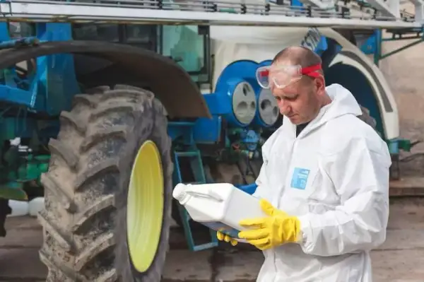 Rolnik w kombinezonie ochronnym i rękawicach trzyma butelkę z preparatem, prawdopodobnie zawierającym difenokonazol, obok traktora.